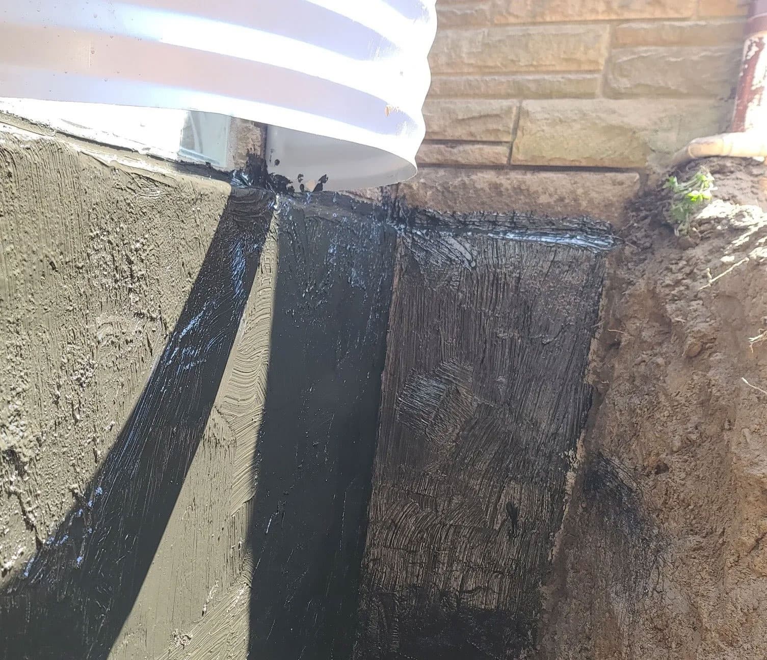Exterior foundation wall coated with black rubberized waterproofing membrane behind a downspout
