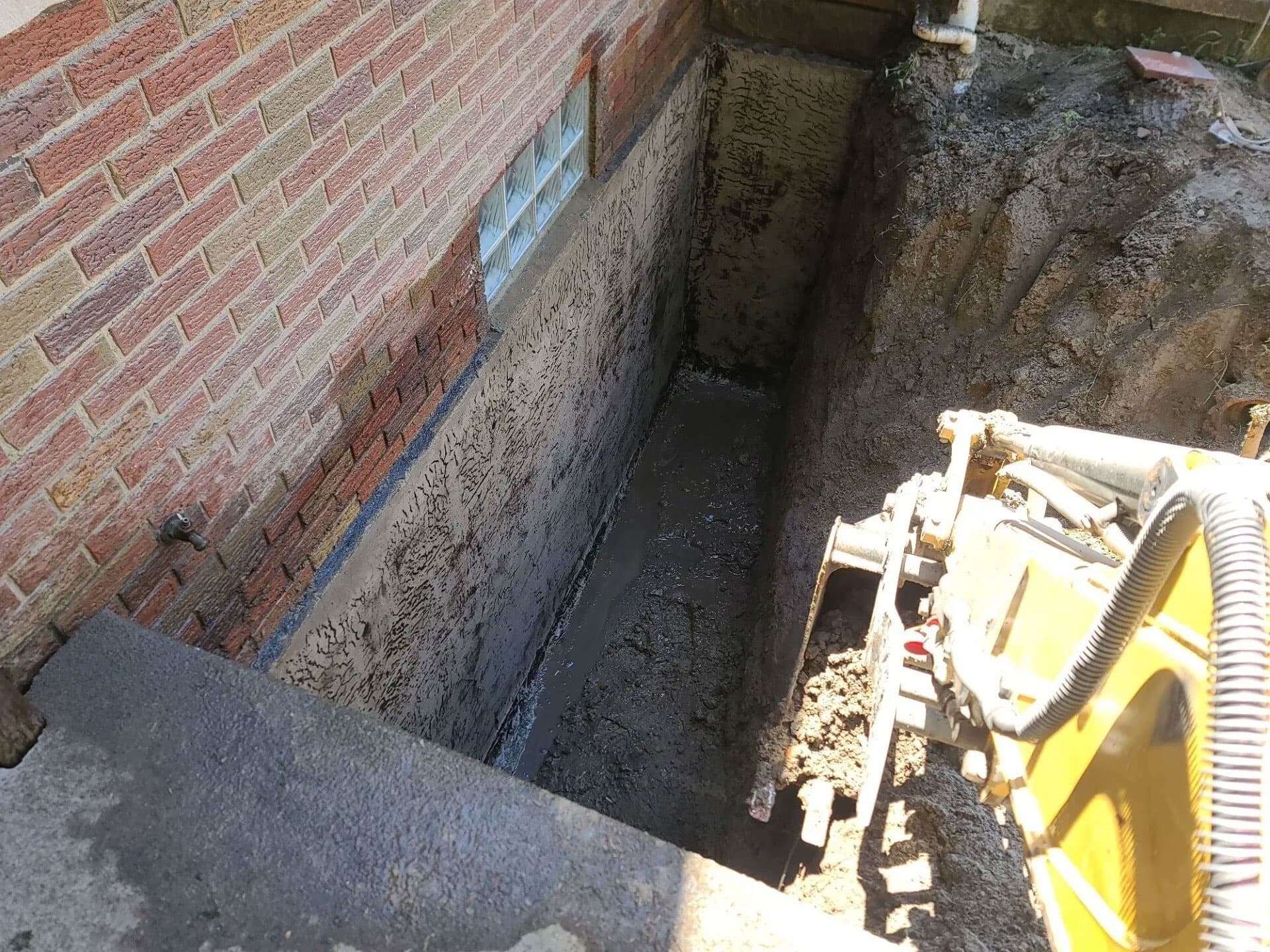 Mini excavator working in a deep trench next to a brick home foundation